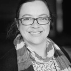 The image is a black and white picture of a women smiling to the camera wearing glasses and a scarf.