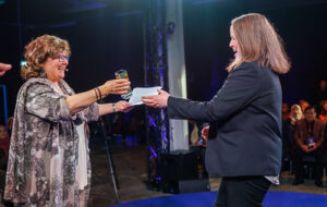 Two women stand on a stage facing side on to the camera. One is handing an award to the other. Both are smiling.