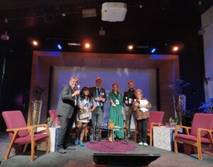 Three women and three men face the camera smiling. They are on a stage holding wooden sticks with marshmallows attached.
