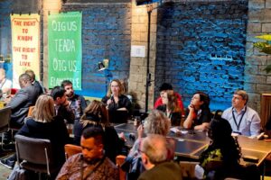 A smaller group of people are seated around a table. A women is speaking into a mircophone.