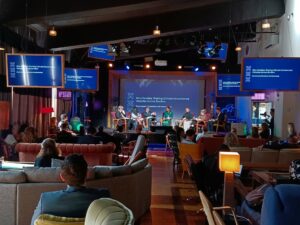 The image is taken from behind the audience. Attendees sit on sofas listening to the 5 speakers on stage.